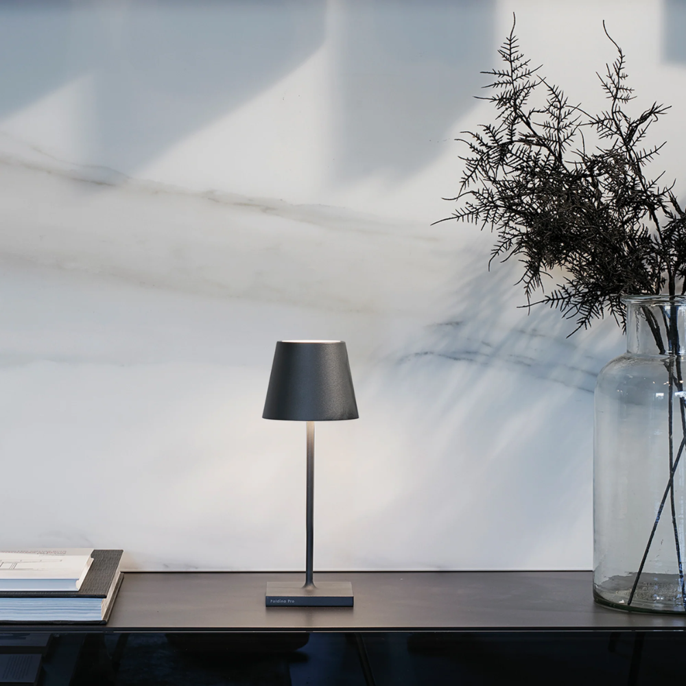 Lampada da tavolo nera in metallo con base quadrata su piano scuro, accanto a libri e vaso di vetro con rami secchi, sfondo marmo.