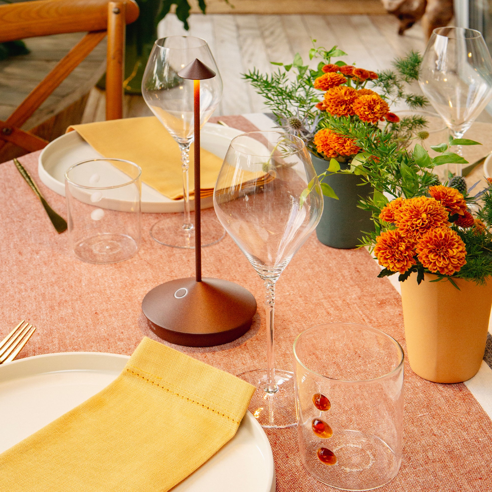 Tavola apparecchiata con tovagliolo giallo, bicchieri trasparenti, fiori arancioni in vaso e lampada metallica marrone.
