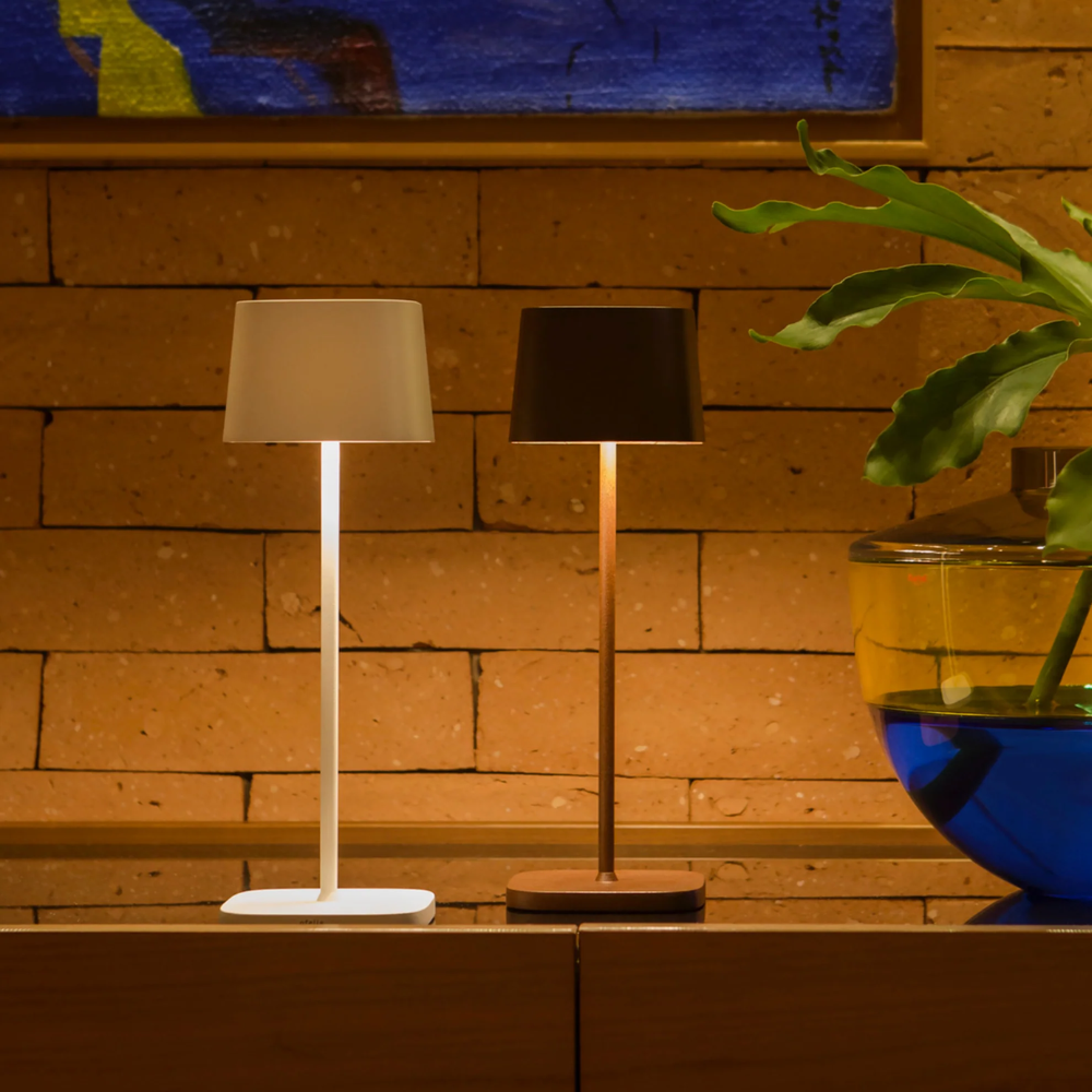 Zwei zylindrische Tischlampen, eine weiße und eine braune, auf Holzschrank vor Steinwand, mit transparenter Vase auf