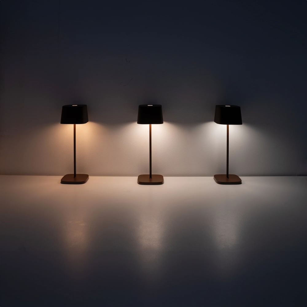 drei schwarze Tischlampen mit dünnem Fuß und Stiel, mit unterschiedlicher Lichtintensität auf weißer Oberfläche und dunklem Hintergrund