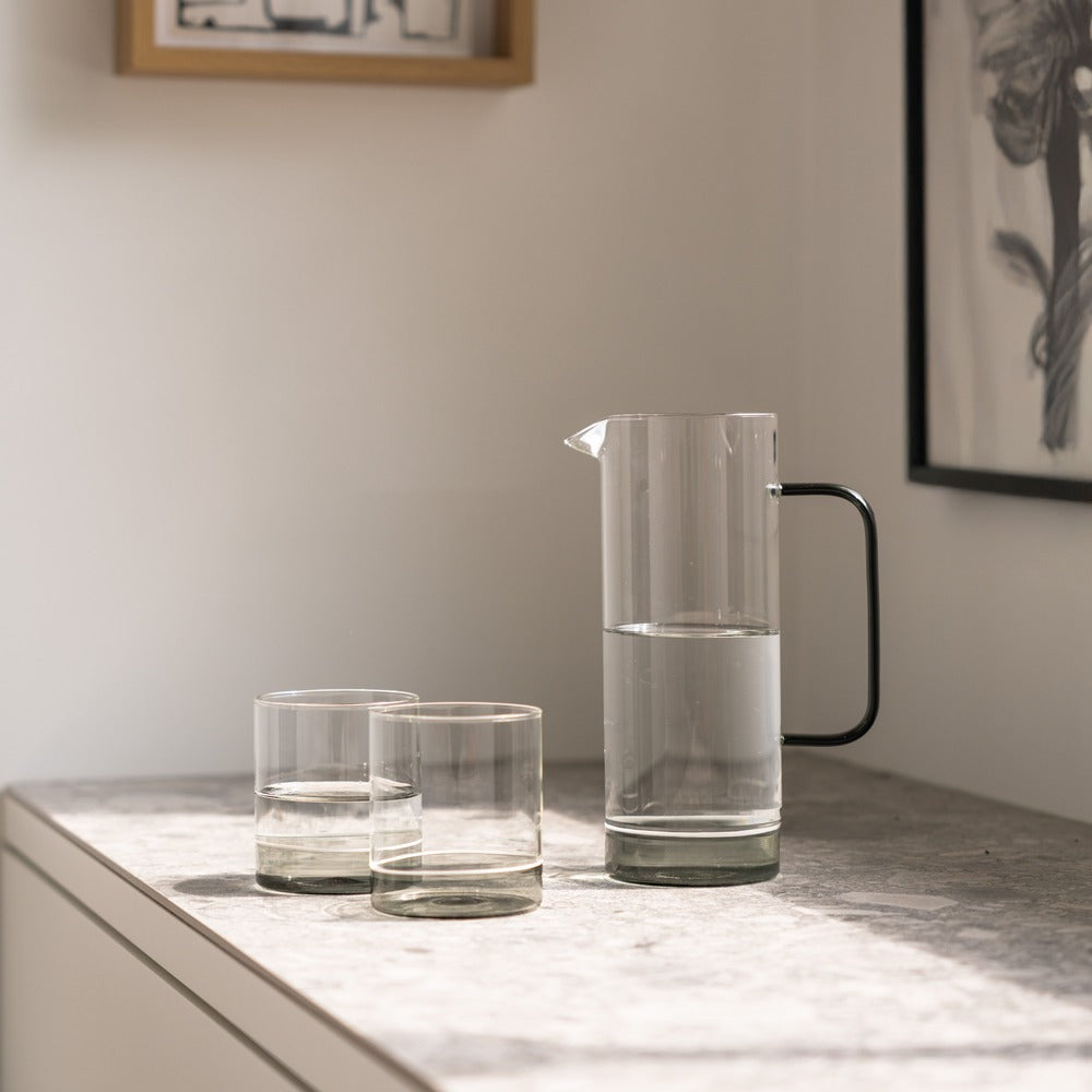 Garrafa de vidrio transparente con asa negra, dos vasos al lado, sobre una mesa de superficie clara.