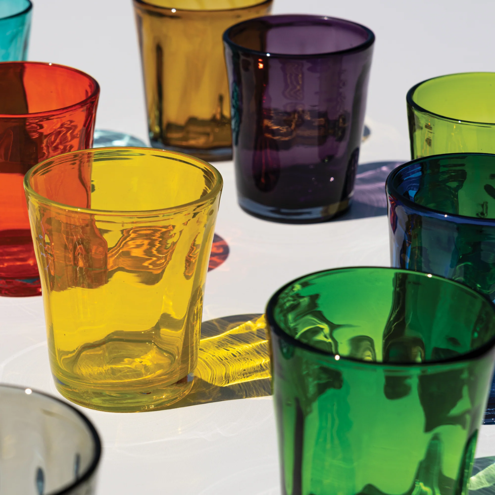 Transparante glazen in diverse kleuren waaronder geel, groen, oranje, blauw en paars op wit oppervlak met gekleurde schaduwen
