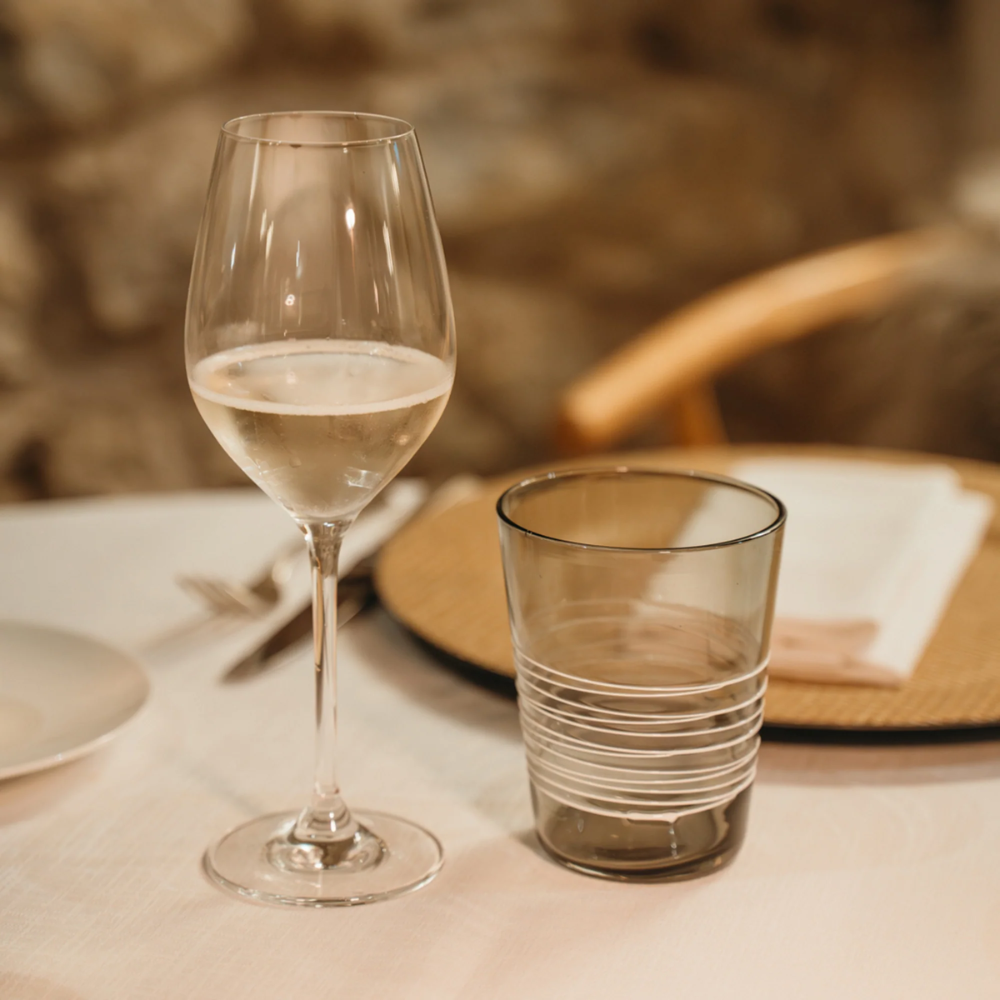 Bicchiere da vino trasparente riempito a metà e bicchiere di vetro fumé con righe bianche su tovagliato bianco, sfondo sfocato.