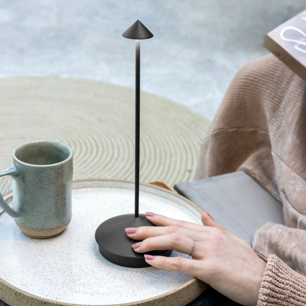 Supporto nero opaco con base tonda e asta sottile verticale con cappello conico, su tavolino con tazza ceramica grigia.