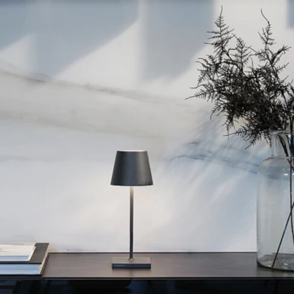 Lampada da tavolo nera con paralume conico, base rettangolare, su superficie scura con libri e vaso trasparente con ramo secco, sfondo