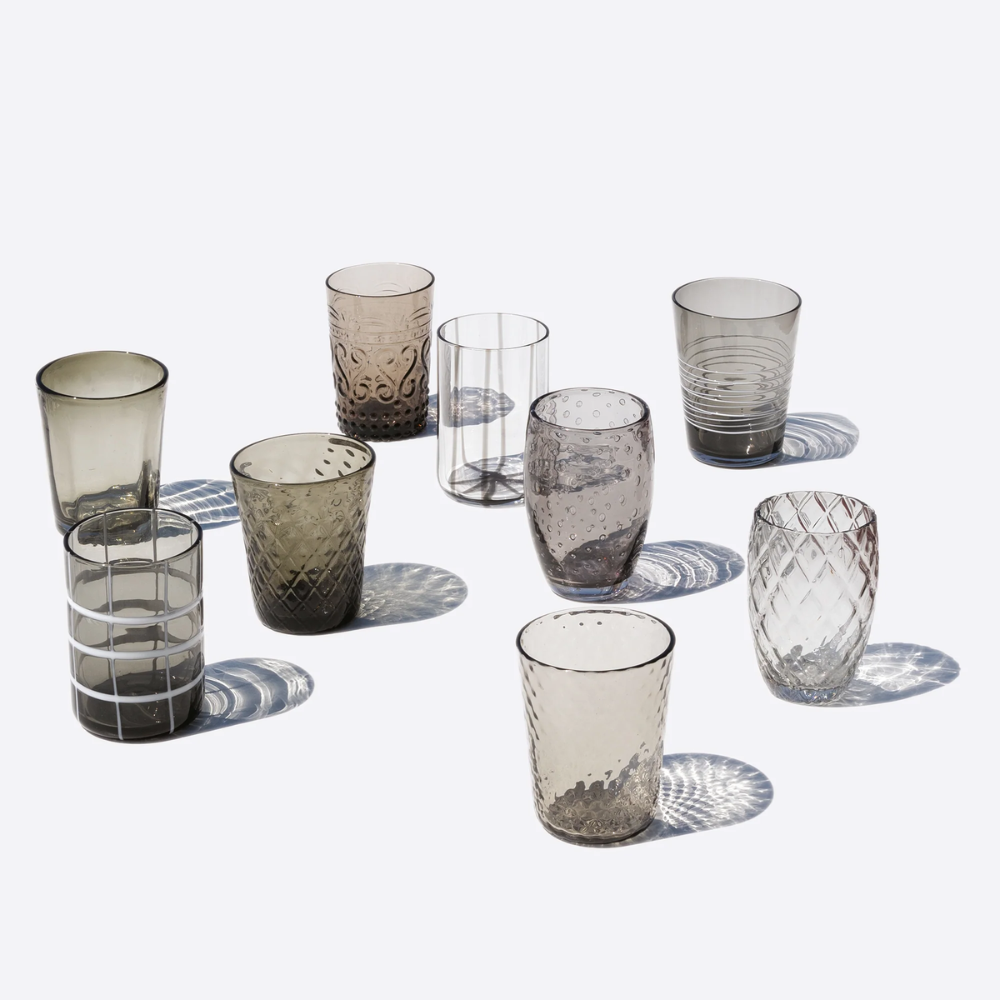Nove bicchieri di vetro trasparente e fumé con diverse texture decorate, su sfondo bianco con ombre nette.