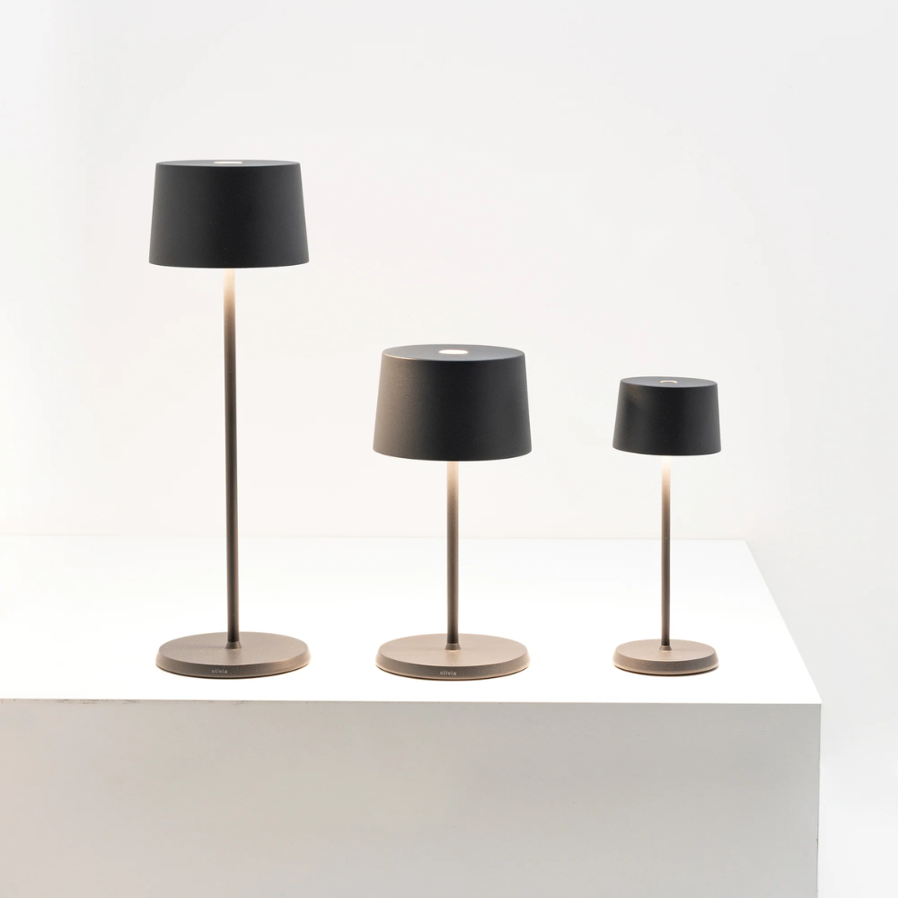 Trois lampes de table modernes avec base et tige couleur bronze et abat-jour noir, de tailles différentes sur surface blanche.