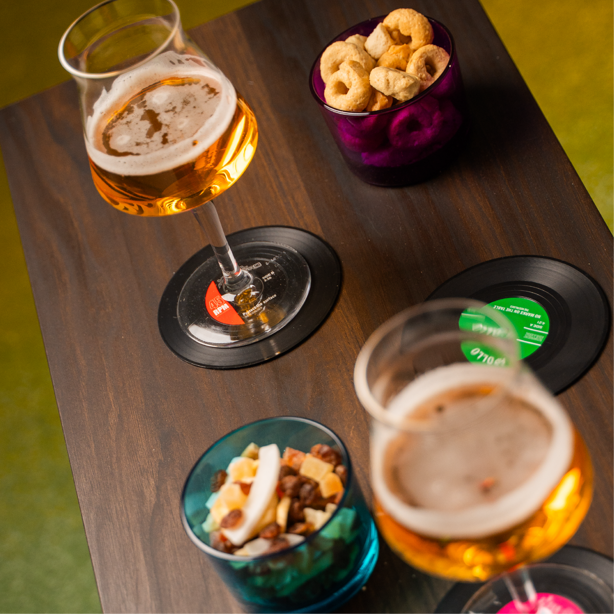 due calici di birra dorata su sottobicchieri a forma di vinile e ciotole con snack su tavolo in legno scuro