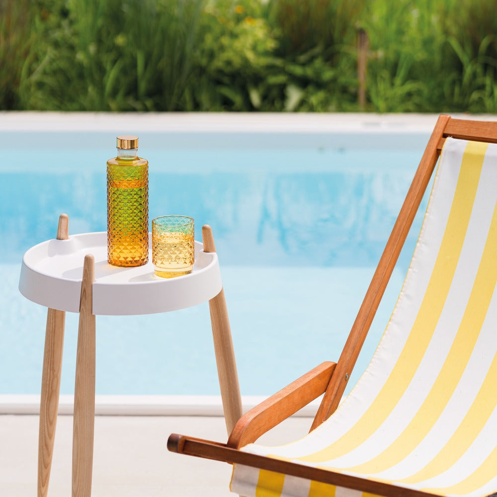 Carafe et verre colorés sur table blanche, à côté d'une chaise longue en bois à rayures jaunes et blanches, près de la piscine.