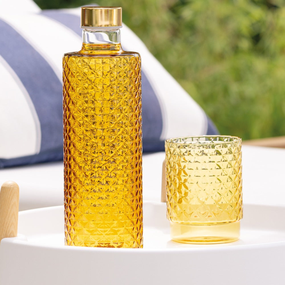 Bouteille et verre en verre transparent, facettés, de couleur jaune ; fond avec coussins bleu et blanc.