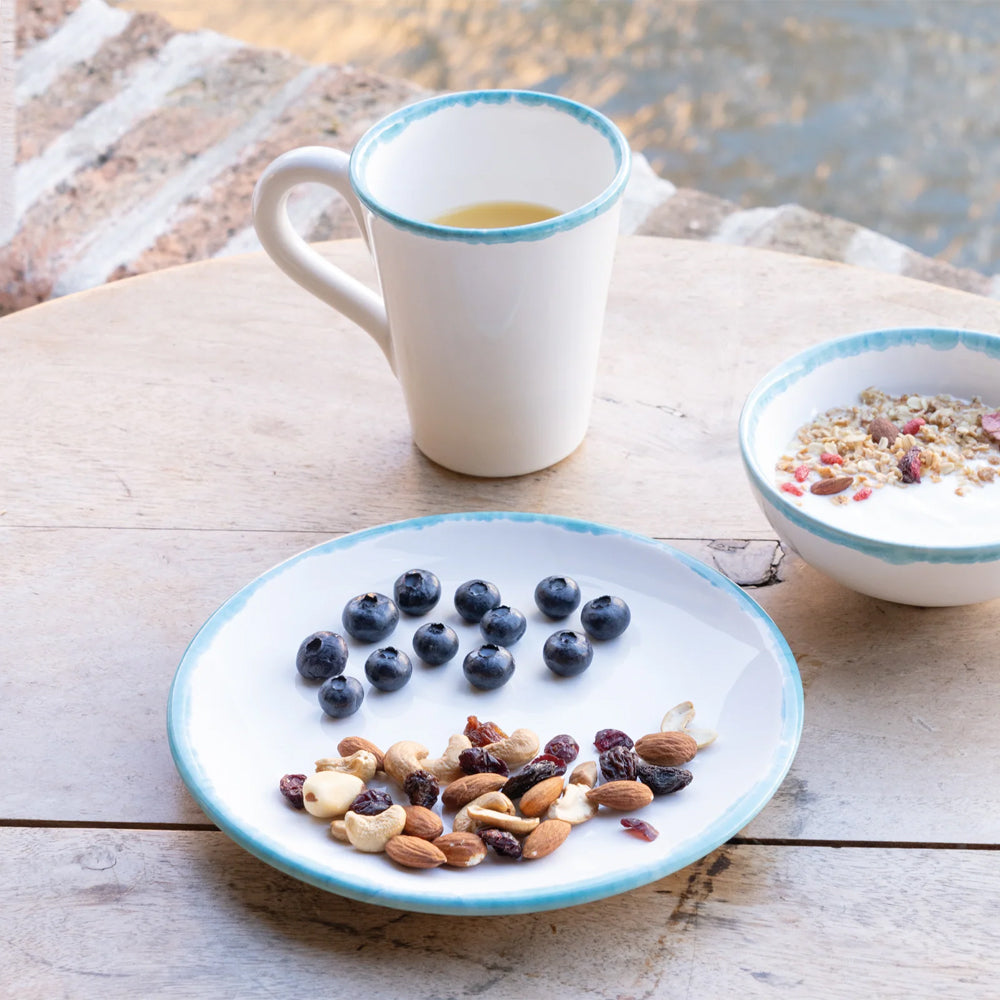 Bord met gedroogd fruit en bosbessen op houten tafel, witte kop met blauwe rand en kom yoghurt met granen.