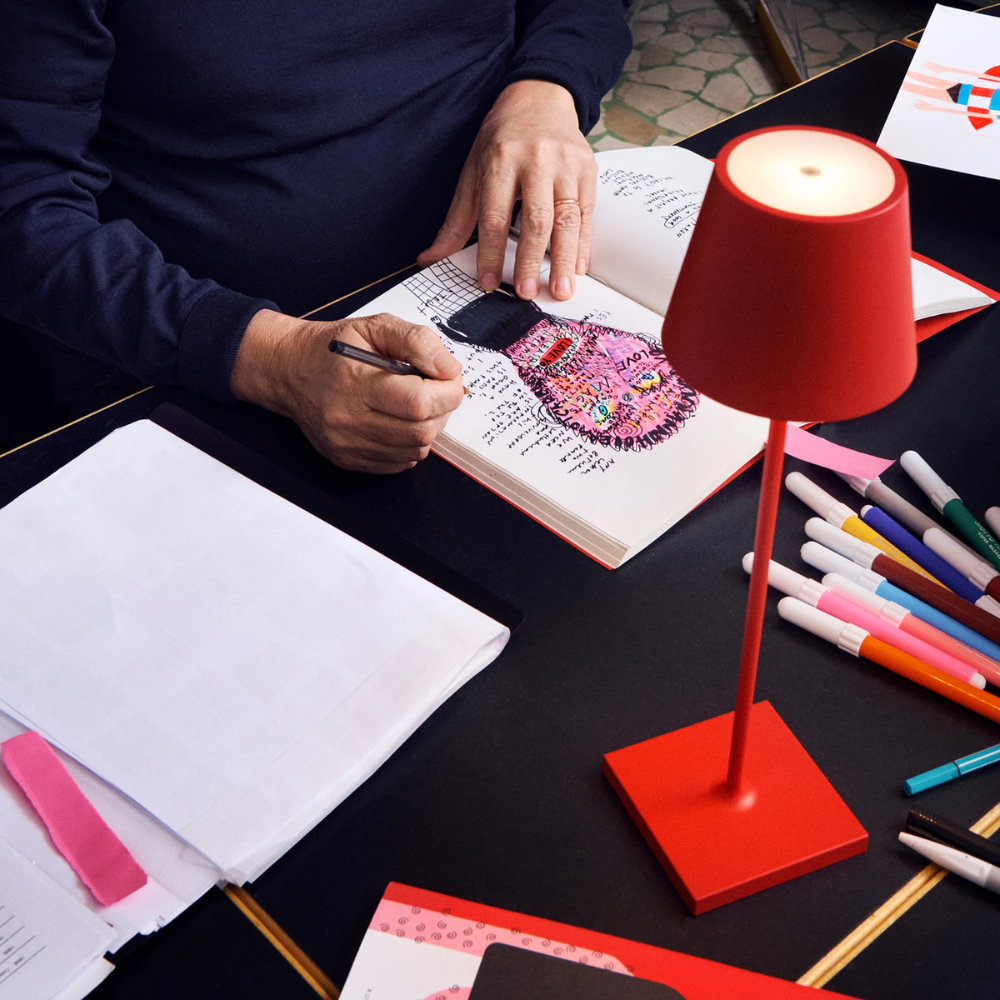Persona che disegna con pennarello su un quaderno con illustrazione colorata, accanto lampada da tavolo rossa e pennarelli vari.