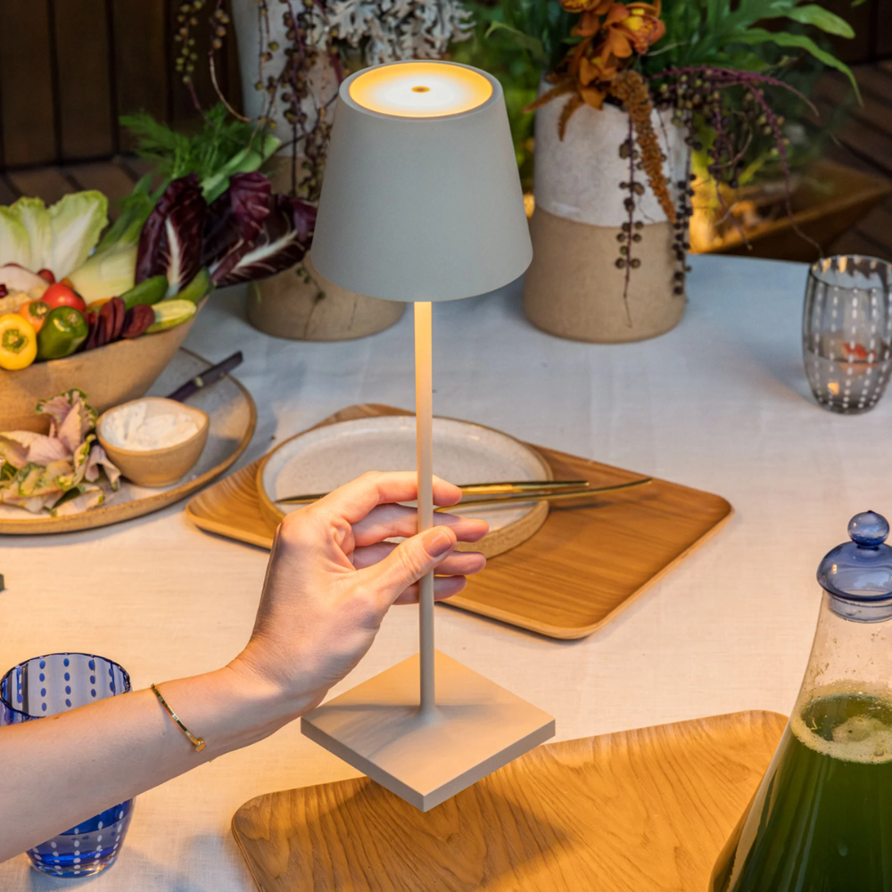 Lampada da tavolo beige in metallo con paralume conico, tenuta a mano su tavola apparecchiata con piatti, bicchieri e decorazioni floreali.