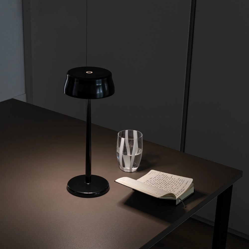 Lampe de table noire brillante avec base et piédestal ronds, verre transparent avec eau et cahier ouvert écrit sur table sombre