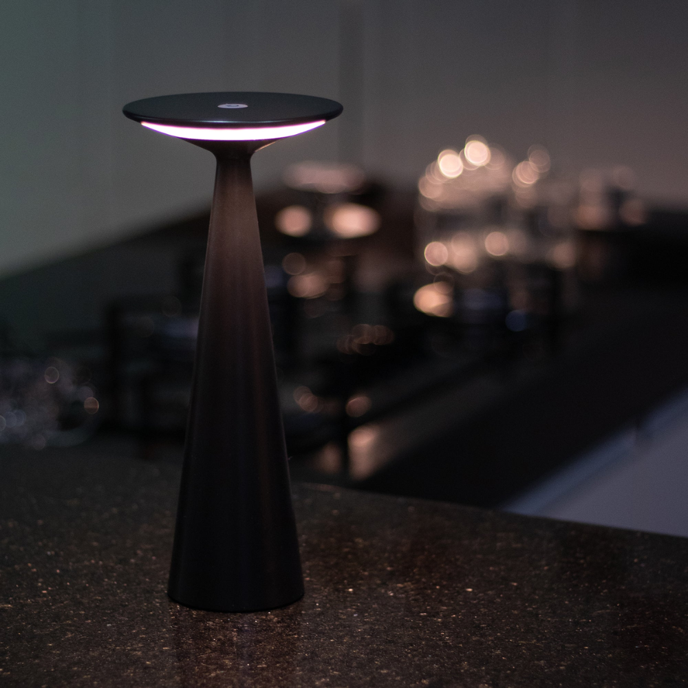 Schwarze kegelförmige Lampe mit breiter Basis und flacher Spitze, beleuchtet mit weißem Licht, auf dunkler Fläche in unscharfer Küche auf dem