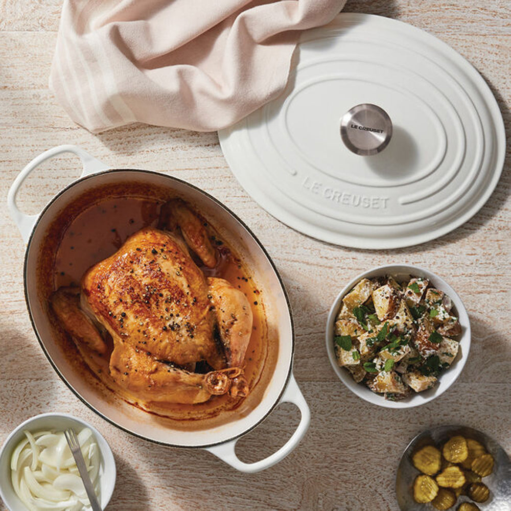 Poulet rôti doré dans une cocotte ovale blanche en céramique avec couvercle Le Creuset, accompagnements de pommes de terre, oignons et concombres dans des bols sur une table