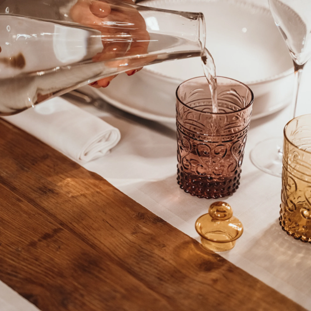Hand schenkt water uit transparante karaf in paars glas met gegraveerde decoratie, wit tafellaken en lichte houten tafel