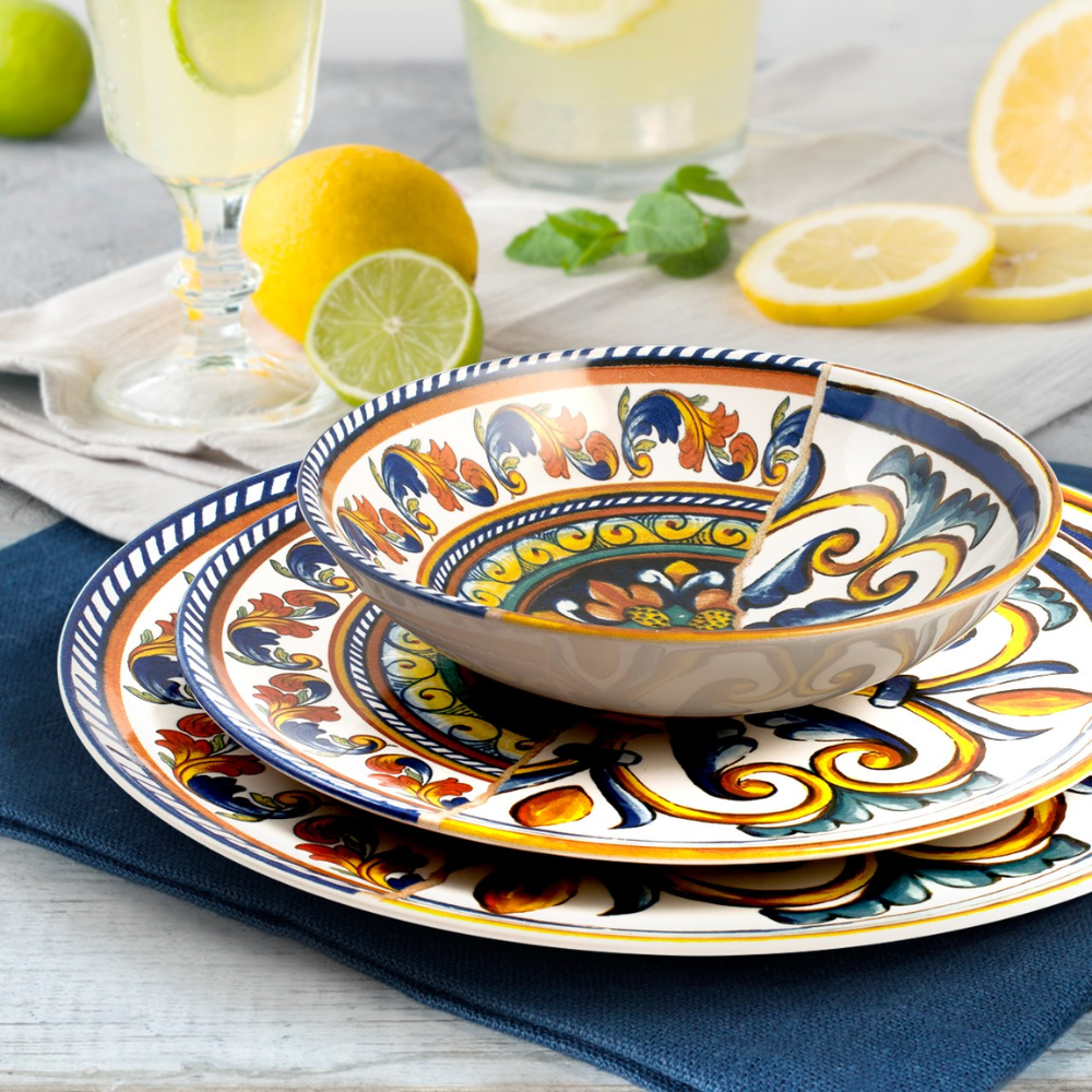 Set da tavola in ceramica decorata con motivi floreali e arabeschi in giallo, blu e arancione sovrapposta su tovagliolo blu con limoni e