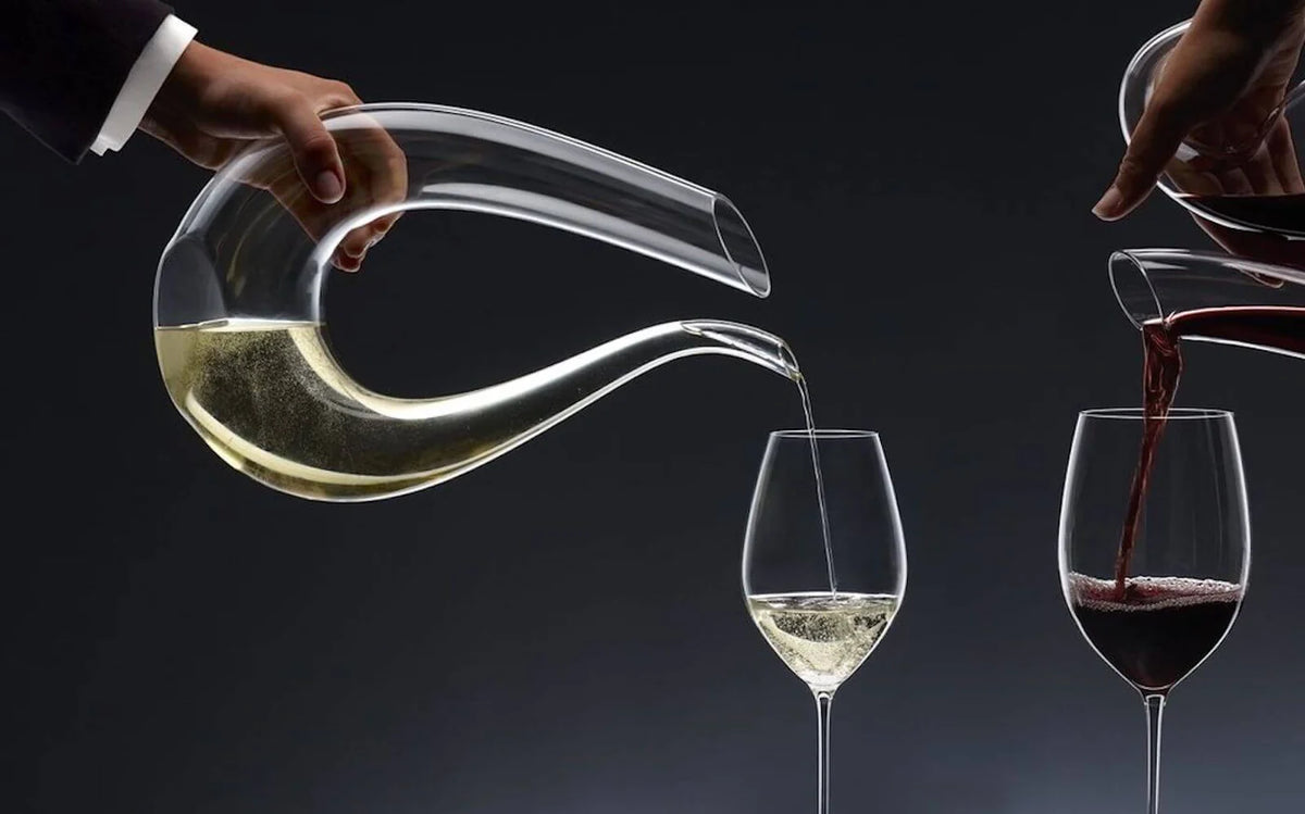 decantatore di vetro trasparente a forma sinuosa e due calici con vino bianco e rosso versati da mani su