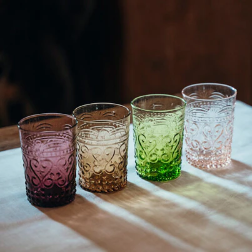 Vier gekleurde geëtste glazen met decoratieve motieven, van links paars, amber, groen en roze, op wit tafelkleed.