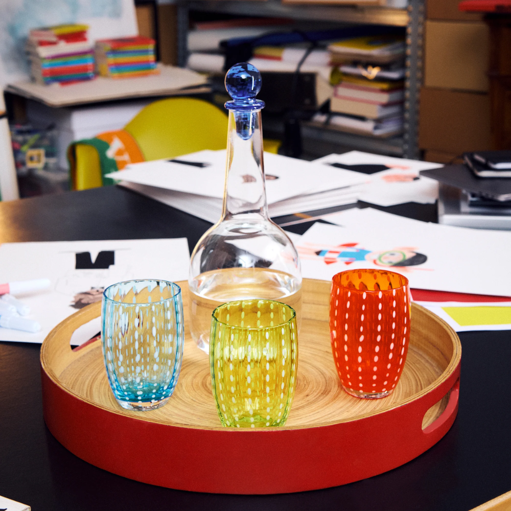 Rond houten dienblad met rode rand met transparante glazen fles en drie gekleurde stippenglazen, op tafel met papieren en boeken.