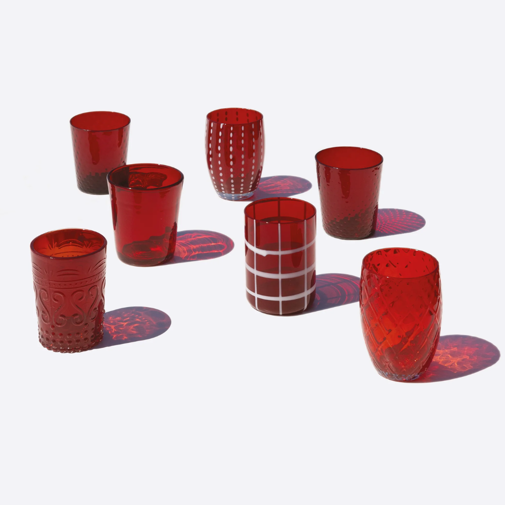 Set di sette bicchieri rossi di vetro con varie decorazioni e texture, disposti su fondo bianco con ombre nette.