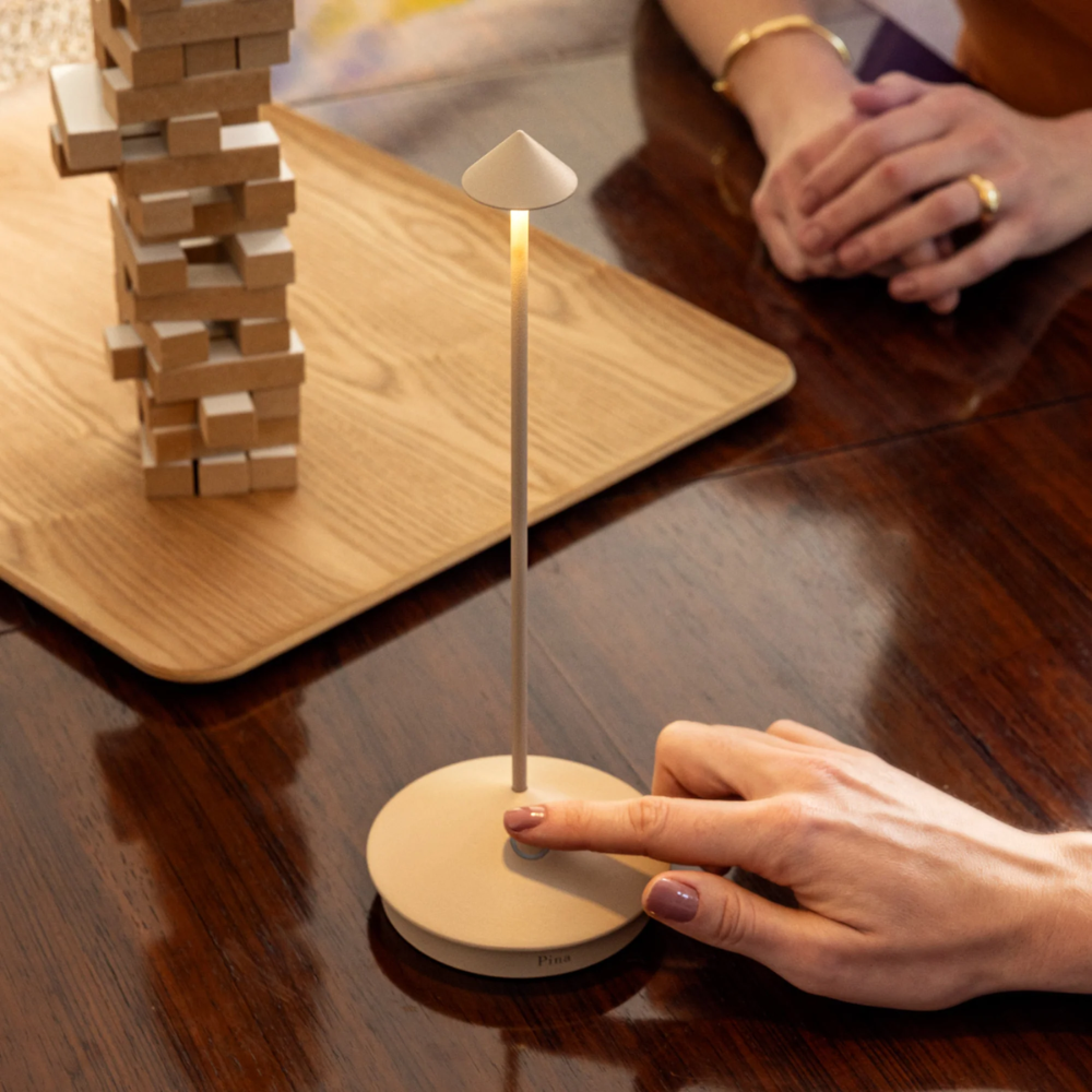 Lampada da tavolo beige con base circolare piatta e paralume conico, su tavolo legno scuro con mani e gioco di blocchi in legno chiaro