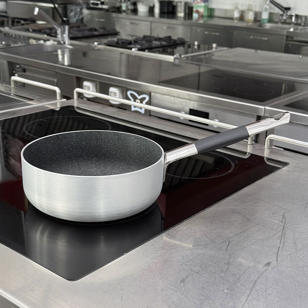 Geborstelde metalen pan met zwarte anti-aanbakken binnenkant en lange stalen steel met zwart gedeelte, op zwarte inductiekookplaat