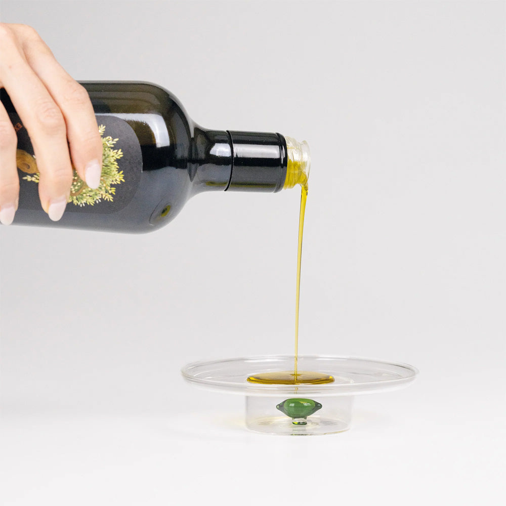Bottiglia nera di olio d'oliva, versata su un piatto trasparente, con olio giallo brillante che scorre, sfondo grigio chiaro.