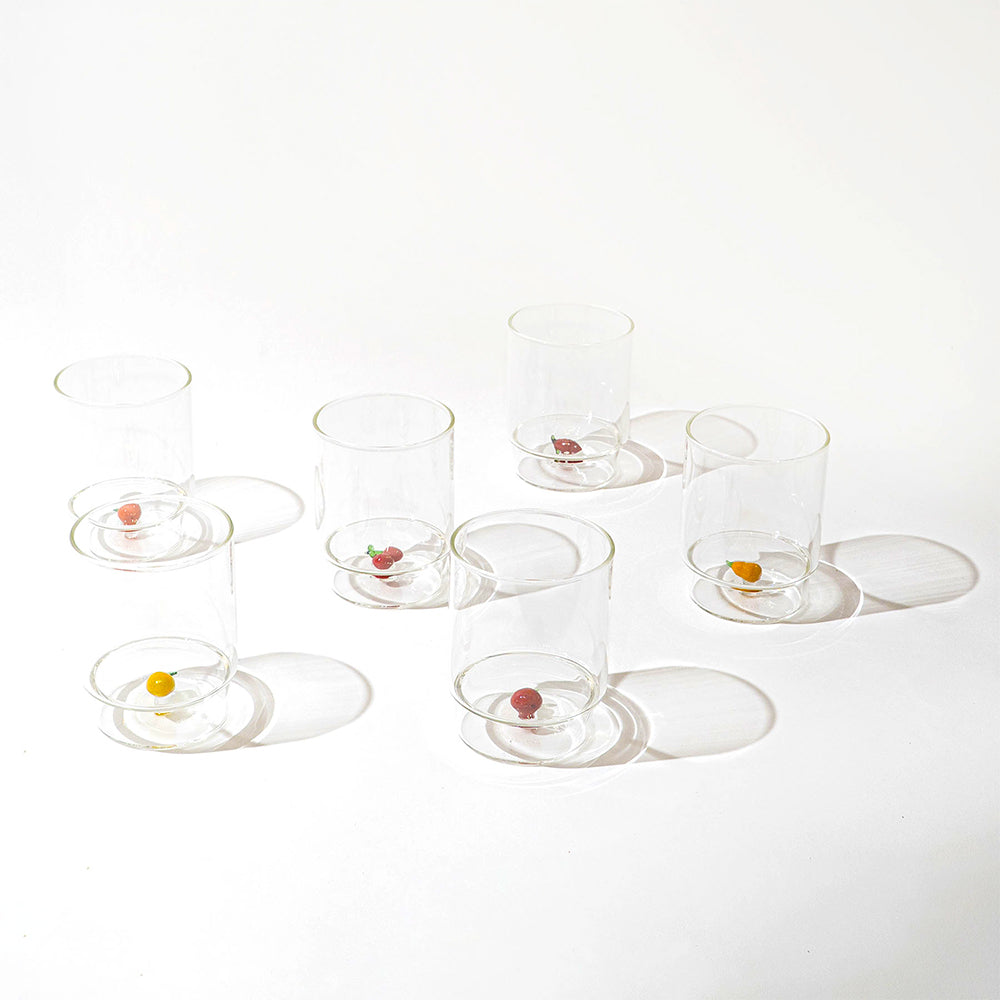 Set di sei bicchieri trasparenti di vetro con piccoli oggetti colorati alla base, disposti su superficie bianca con ombre nette.