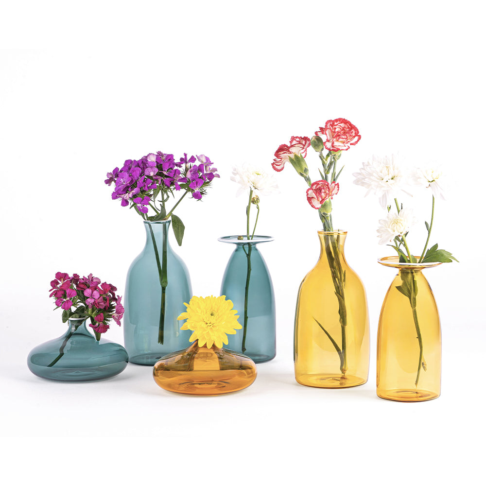six vases en verre transparent, trois bleus et trois jaunes, avec fleurs de diverses couleurs insérées, sur fond blanc