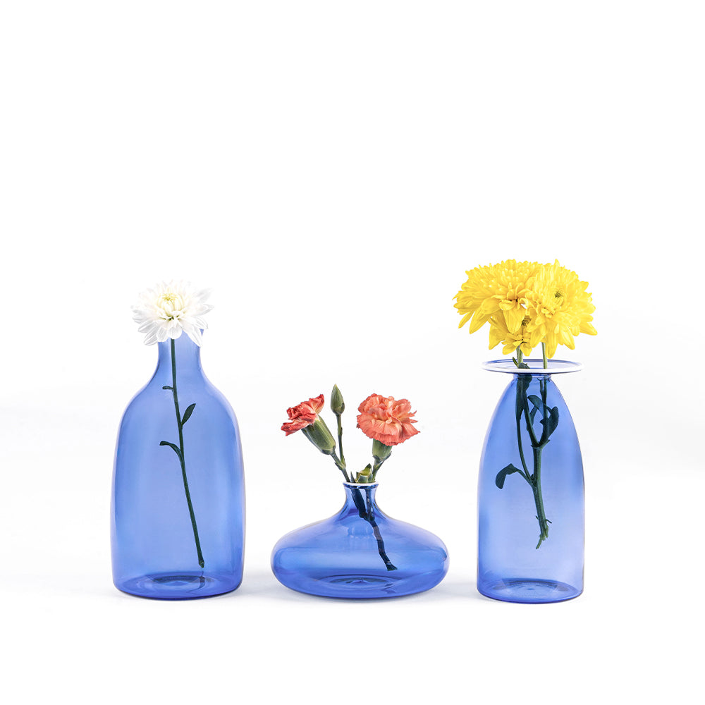 Trois vases bleus transparents en verre de formes différentes avec fleurs fraîches blanches, orange et jaunes sur fond blanc.