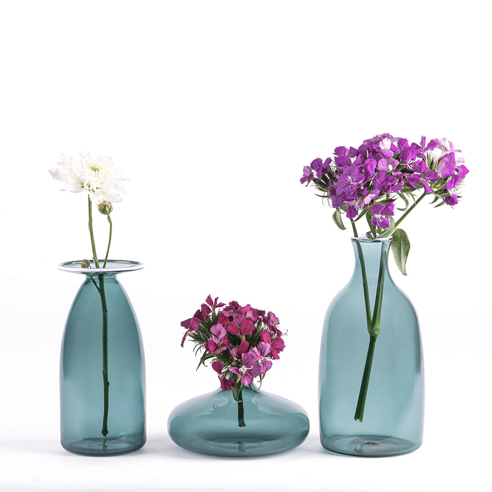Trois vases bleu clair en verre transparent avec fleurs blanches et violettes, sur fond blanc minimaliste.