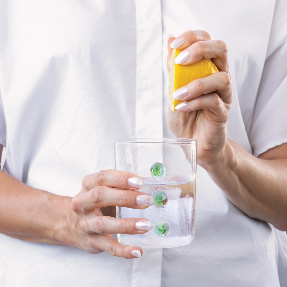 Mains avec ongles vernis blanc tenant un verre en verre avec glace et pressant une tranche de citron jaune sur fond blanc.