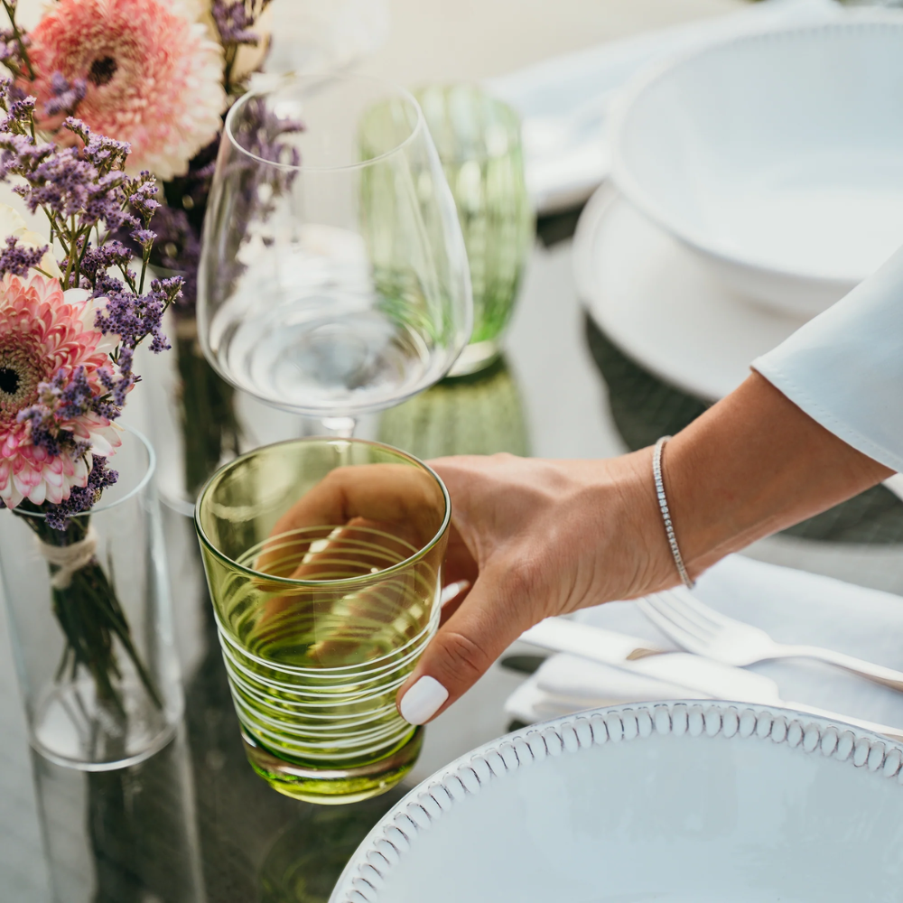 Bicchiere di vetro verde con motivo a righe tenuto da mano femminile, accanto a piatto bianco, coltello, forchetta e vaso con fiori rosa e