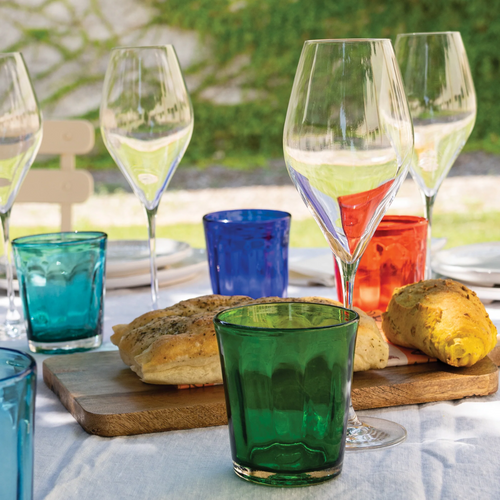 Verre vert en verre épais devant planche avec pain, calices transparents et autres verres colorés, table en extérieur.