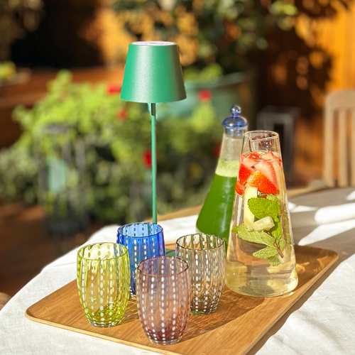 Set di quattro bicchieri di vetro colorati con decorazioni a pallini bianchi, caraffa con acqua, fragole e menta su vassoio di legno.