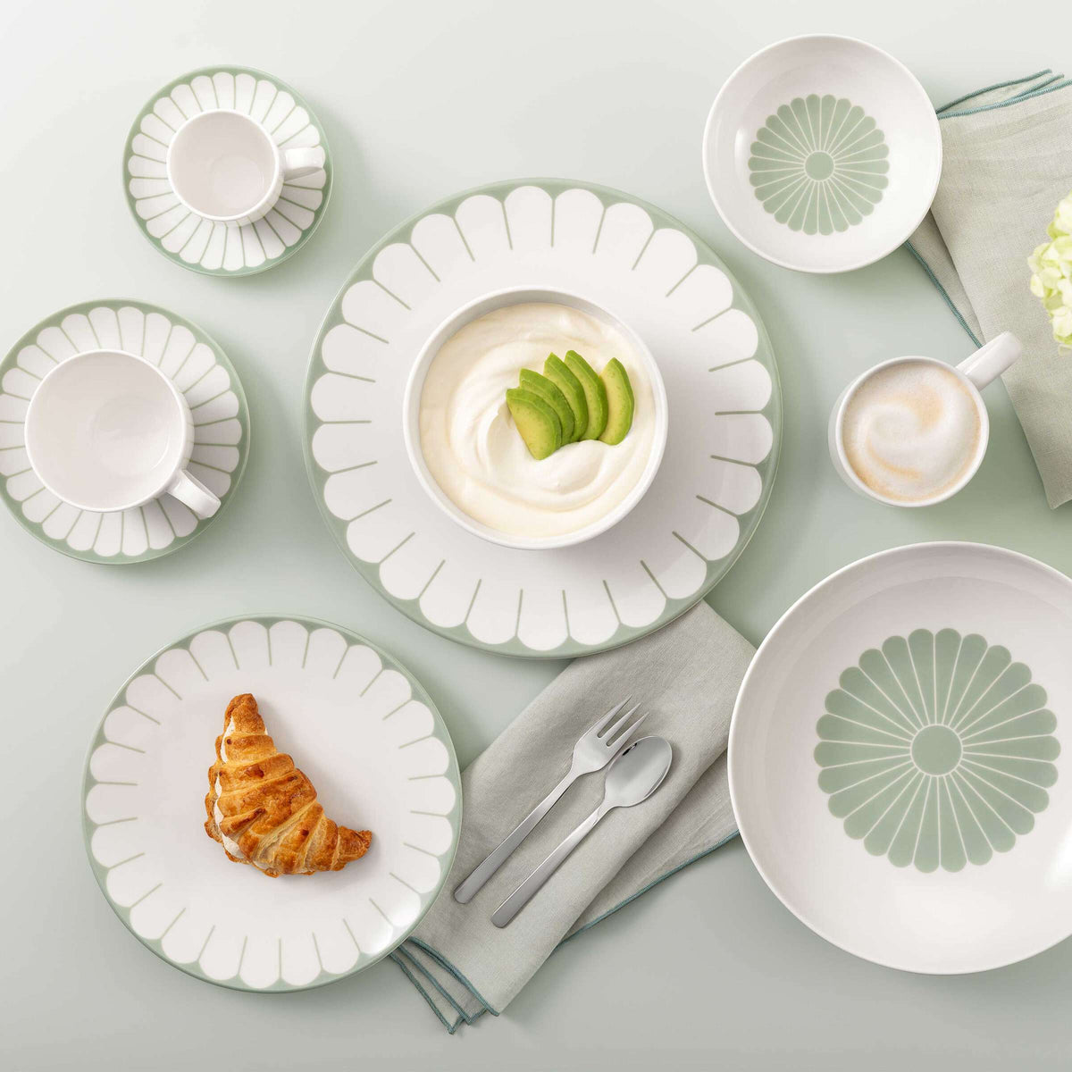 Set di stoviglie bianche con decorazione a raggi verde salvia, con croissant, ciotola con crema e avocado, tazza di caffè e posate su tovagliolo