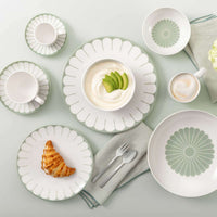 Set da colazione con piatti bianchi decorati, ciotola con yogurt e avocado, croissant su piatto, posate e tovagliolo grigio.