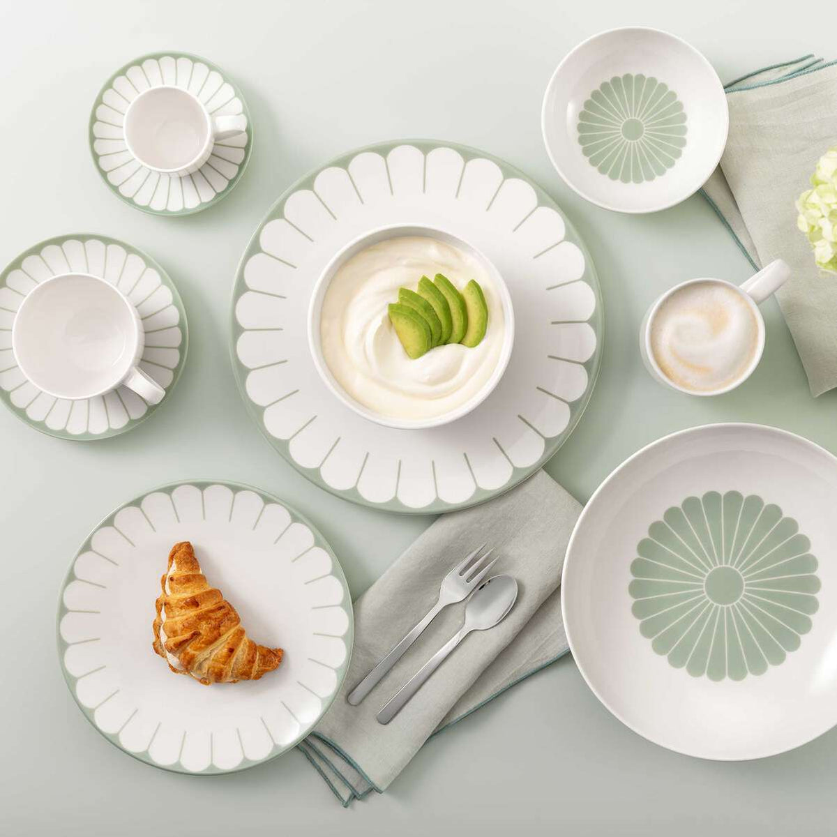 Servizio di piatti e tazze in ceramica bianca con decoro verde a fiore, con cappuccino, yogurt con avocado e croissant su tovagliolo grigio.