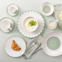 Servizio di piatti e tazze in ceramica bianca con decoro verde a fiore, con cappuccino, yogurt con avocado e croissant su tovagliolo grigio.