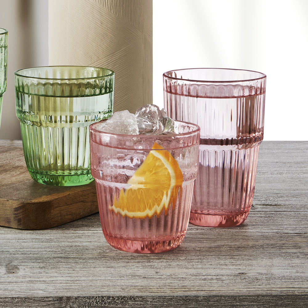 Tre bicchieri di vetro colorato rosa e verde, uno con ghiaccio e fetta d'arancia, su tavolo in legno chiaro
