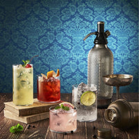 Quattro bicchieri con cocktail colorati su libri e tavolo in legno con seltz in bottiglia metallica traforata e shaker metallico, sfondo