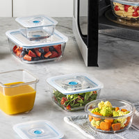 Transparente Glasbehälter mit Deckeln, gefüllt mit Obst, Gemüse und Pürees, auf einer Marmorfläche.