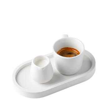 Tazzina bianca con caffè su vassoio ovale bianco insieme a piccola brocca bianca per latte, sfondo bianco.