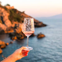 Verre transparent avec gravures blanches contenant du vin rosé, tenu par une main sur fond de mer et falaise au coucher du soleil.