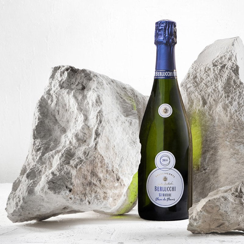 Una botella de vino espumoso Berlucchi, con etiqueta azul, colocada entre rocas claras sobre fondo blanco.