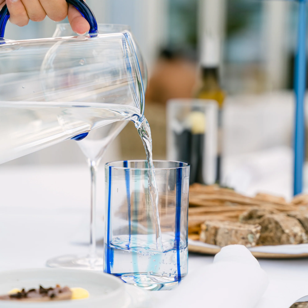 Caraffa trasparente con manico blu versa acqua in bicchiere trasparente con strisce blu, su tavola bianca con piatto di pane e posate.