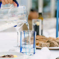 Caraffa trasparente con manico blu versa acqua in bicchiere trasparente con strisce blu, su tavola bianca con piatto di pane e posate.