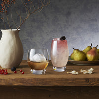 Due bicchieri con bevande, uno con ghiaccio e liquido ambrato, l'altro con ghiaccio e bevanda rosa con mora sopra, vaso beige, pera e